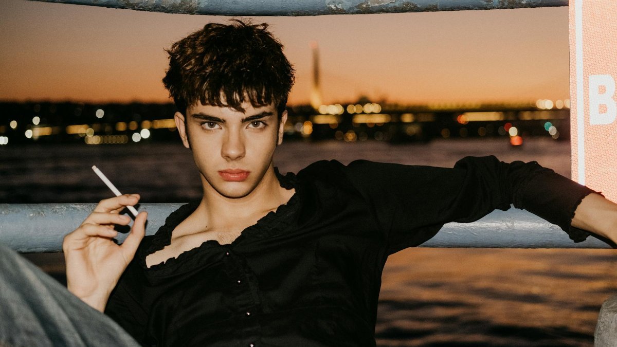 Young male model in black shirt posing by Istanbul waterfront at sunset with city lights.