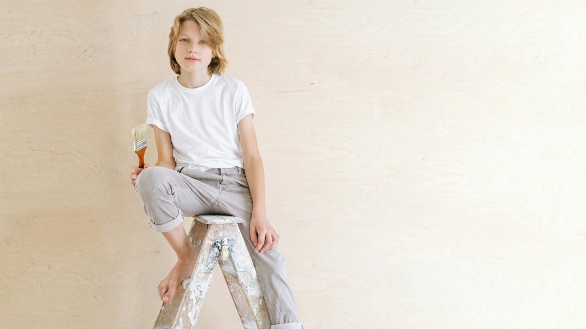 A young boy in casual attire sits on a ladder holding a paintbrush, exuding a creative vibe.