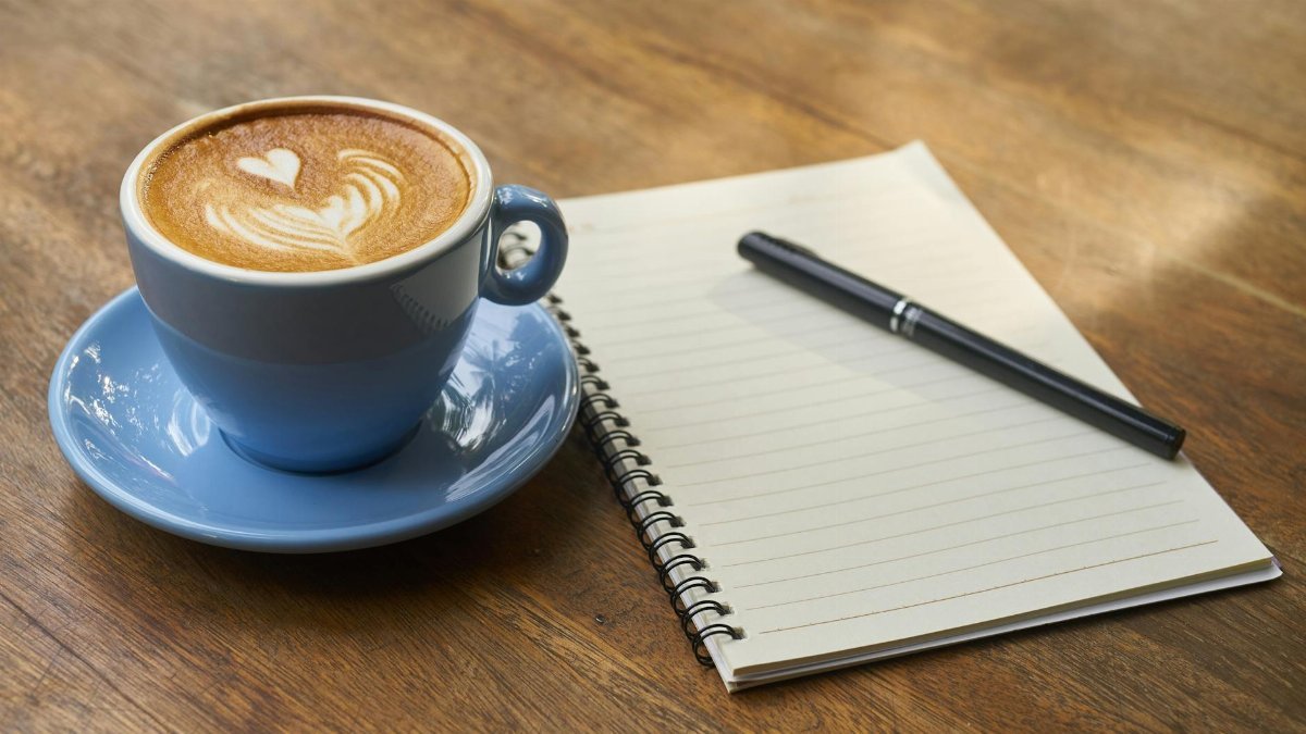 A calming scene of a latte with art in a blue cup beside a notebook and pen on a wooden table.