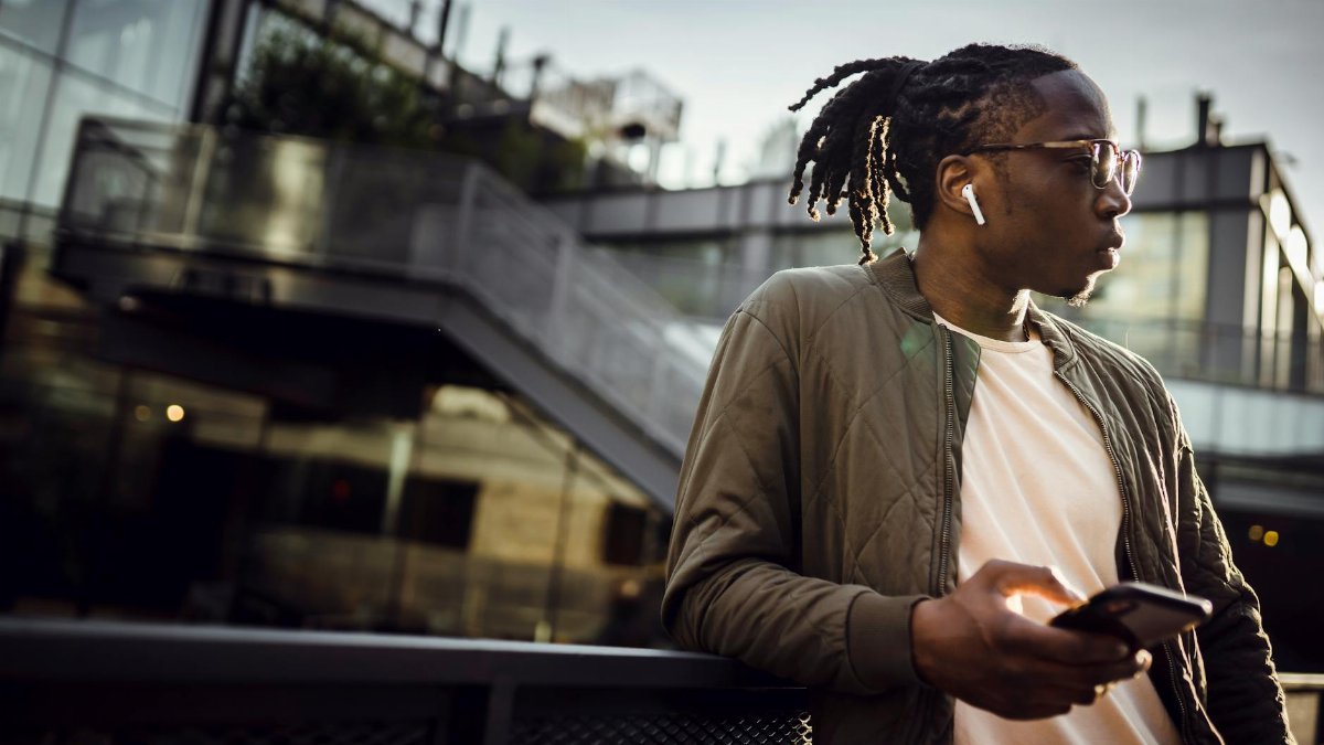 From below of dreamy African American male with pigtails and true wireless earphones standing on bridge with cellphone and listening to music while looking away