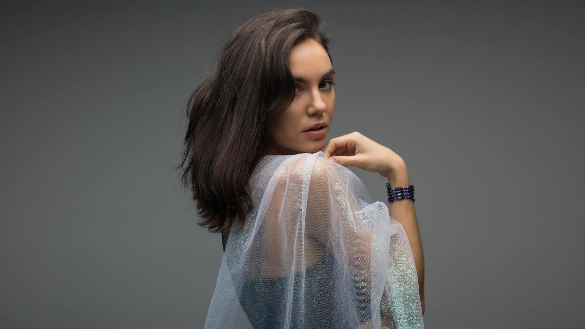 Side view of calm lady covering shoulders with translucent veil and looking at camera while standing in modern studio