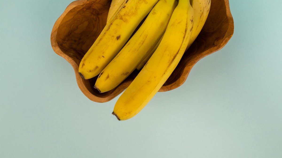 Top view of bunch of fresh bananas with blots on smooth yellow peel in curved bowl on light background