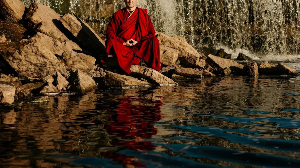 A monk in red robes meditates by a tranquil waterfall, reflecting peace.