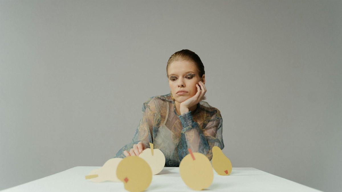 Conceptual portrait of a woman in a studio with paper cut-out fruits on a white table.