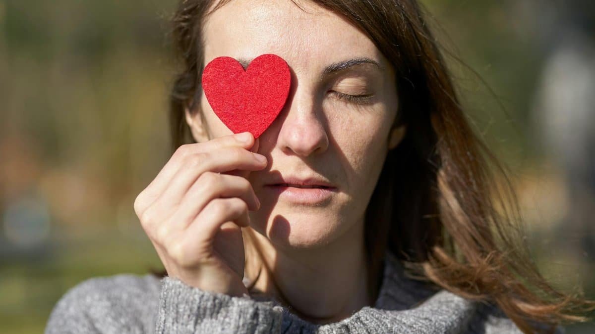 Woman holding a red heart symbol in front of her eye, enjoying a sunny outdoor day.