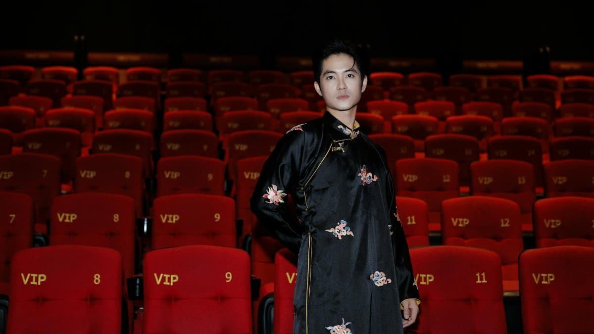 Portrait of an Asian man in traditional clothing standing in a VIP cinema with red seats and floral patterns.
