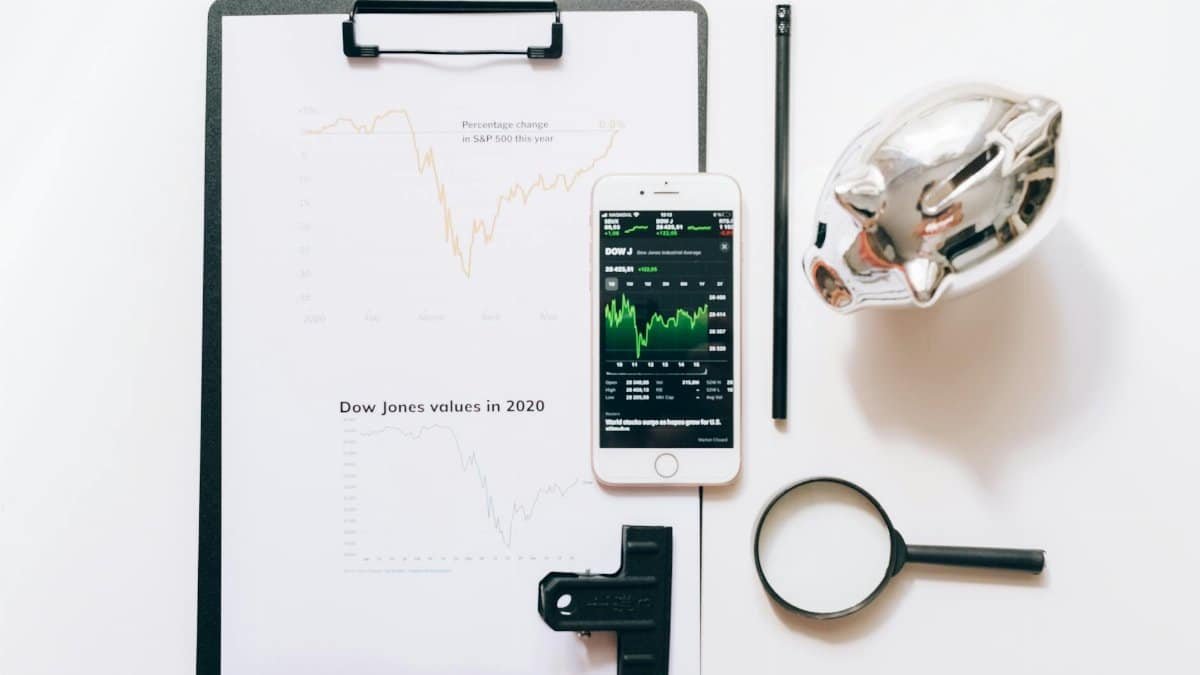 Top view of financial analysis tools including a cellphone, clipboard, magnifying glass, and piggy bank on a white desk.