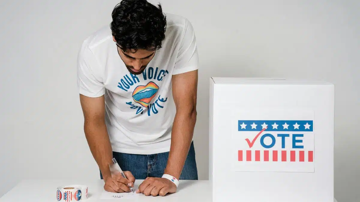 A man writes on a paper to cast his vote next to a ballot box in a studio setting.
