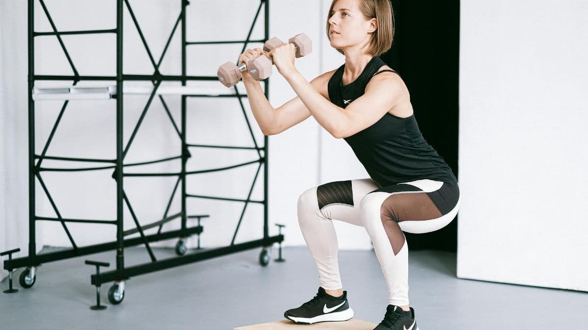 A woman in sportswear performs a squat with dumbbells indoors, embodying strength and healthy living.