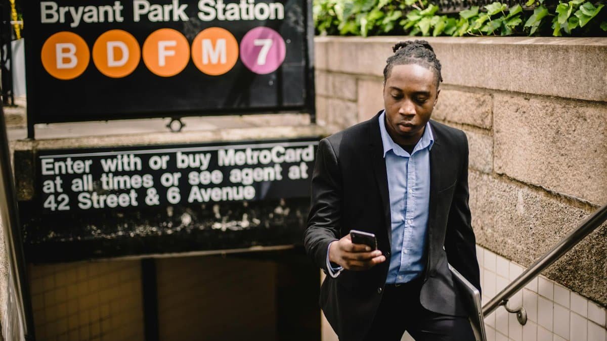 Serious African American office worker with laptop in hand using mobile while exiting subway in Manhattan on sunny summer day