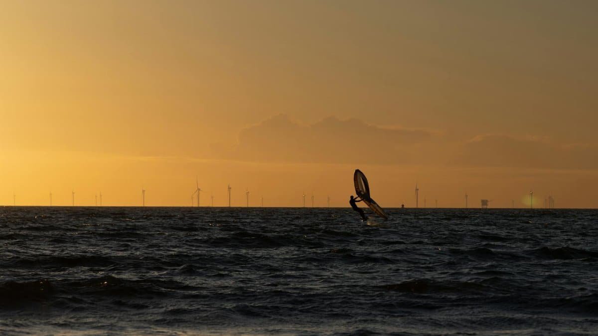Silhouette of a windsurfer against a golden sunset on an open sea, with distant wind turbines in view.