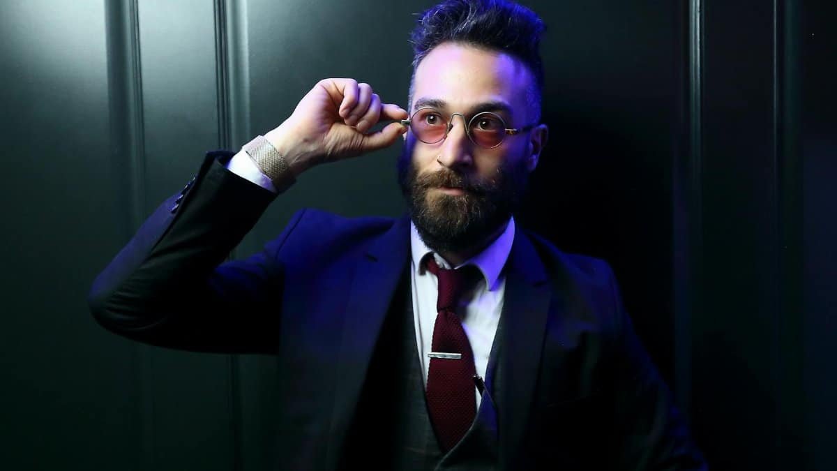 A bearded man with glasses in a stylish suit poses confidently against a dark background, evoking modern sophistication.