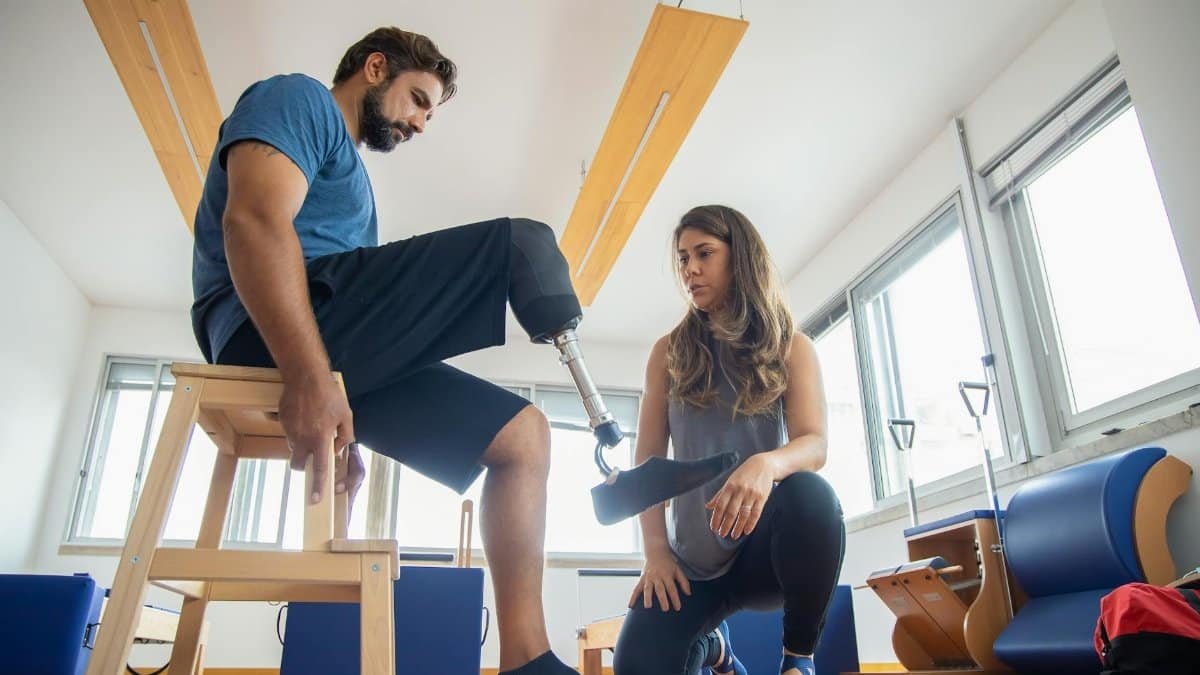 A man with a prosthetic leg assisted by a therapist during rehabilitation indoors.
