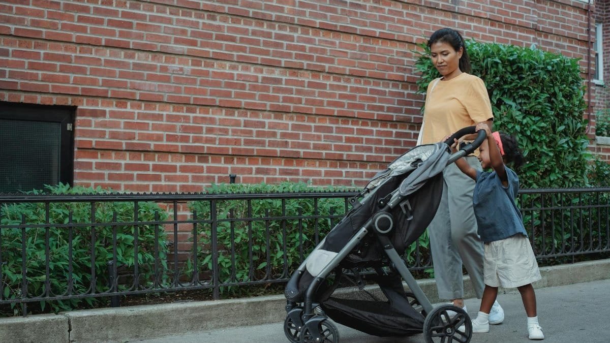 Full length of Asian mom with small girl strolling on footpath with stroller near green hedge in city