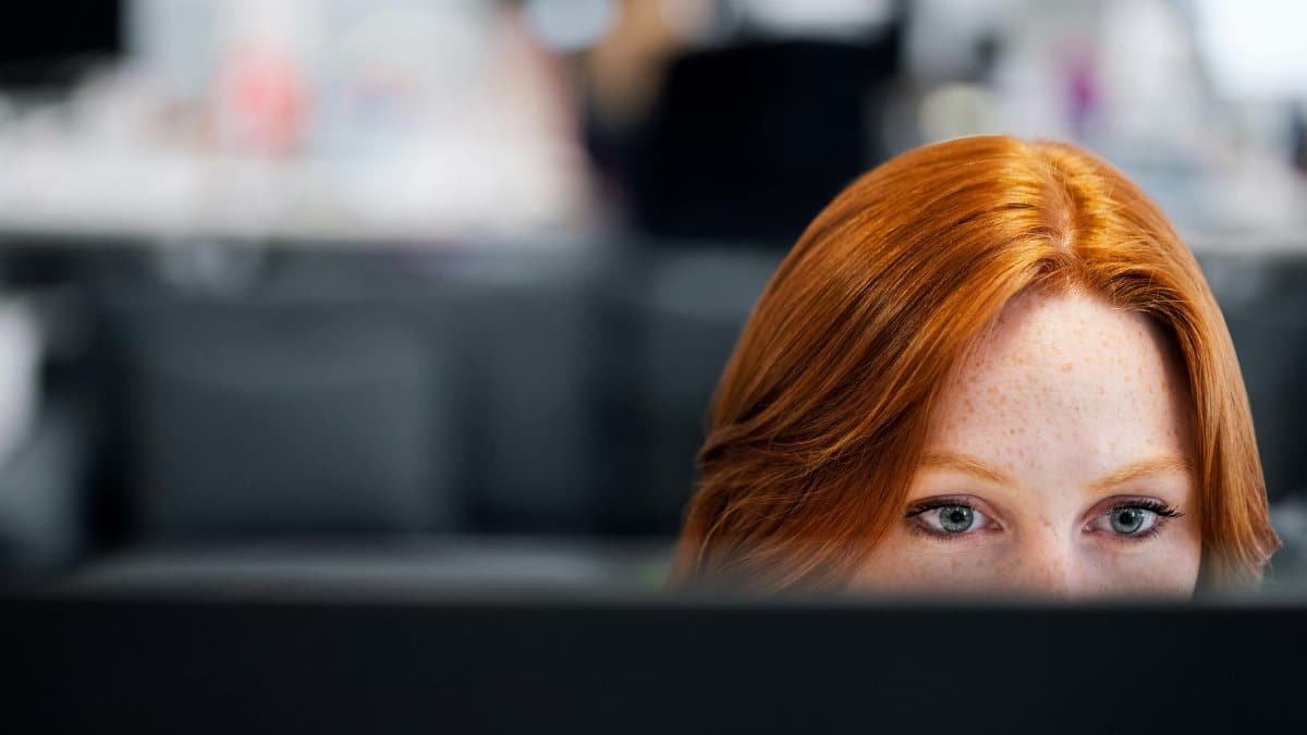 A woman with red hair intensely focused on a computer screen in an office setting.