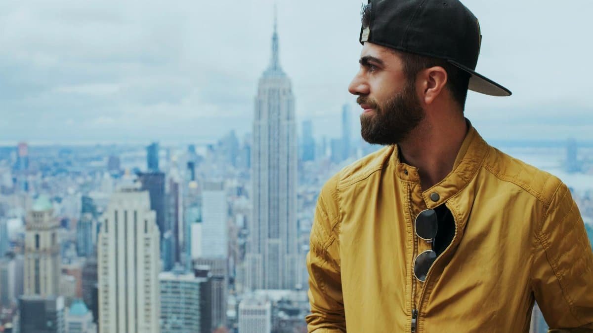 Man with sunglasses and cap stands against New York City's iconic skyline featuring the Empire State Building.