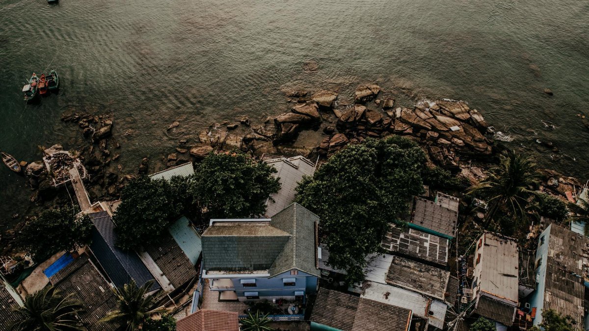 Aerial view capturing a Vietnamese coastal village with houses, trees, and rocky seashore at sunset.