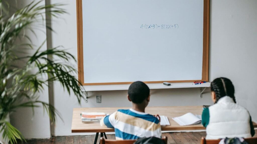 Back view unrecognizable multiethnic pupils sitting at desk with copybooks against whiteboard in light modern classroom