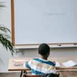 Back view unrecognizable multiethnic pupils sitting at desk with copybooks against whiteboard in light modern classroom