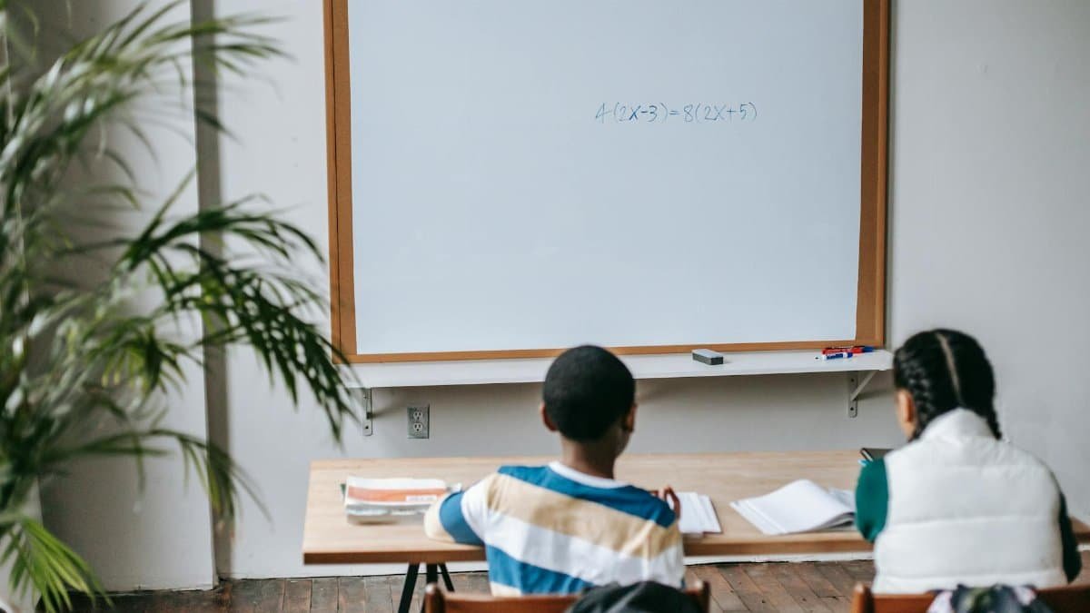 Back view unrecognizable multiethnic pupils sitting at desk with copybooks against whiteboard in light modern classroom