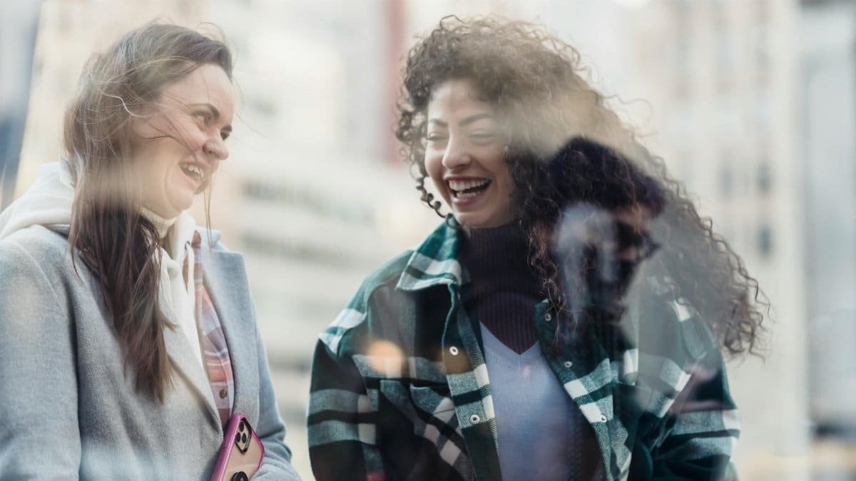 Two women share laughter through a glass window in a city, reflecting joy and friendship.