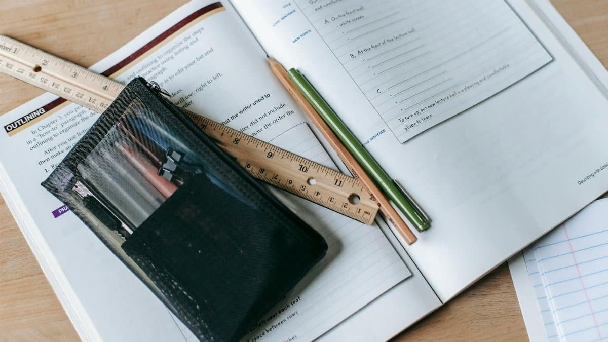 From above of textbook with exercises and ruler with pens near small case on table