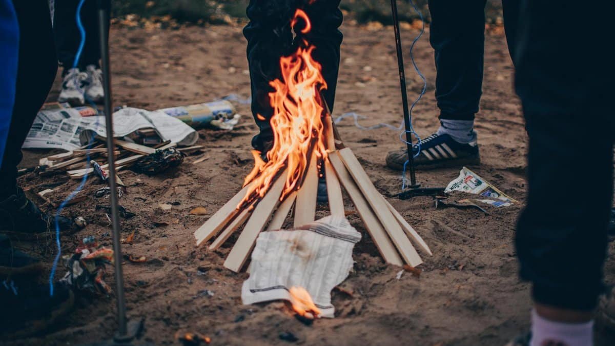A group of people enjoying a campfire outdoors, surrounded by wood and newspapers.