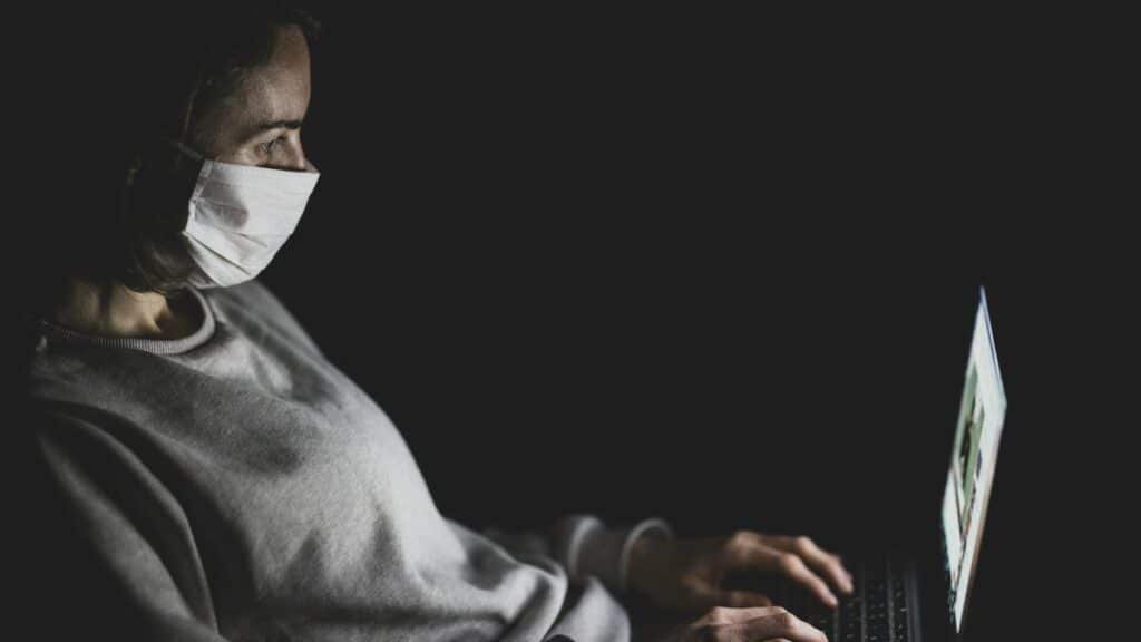 Adult woman wearing a face mask using a laptop in a dark room, emphasizing isolation and digital communication.