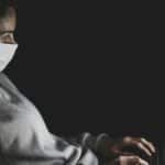 Adult woman wearing a face mask using a laptop in a dark room, emphasizing isolation and digital communication.