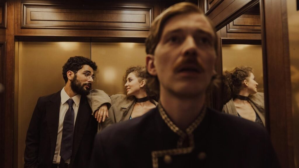 A stylish couple exchanges glances in a wood-paneled elevator alongside a concierge.