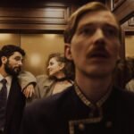 A stylish couple exchanges glances in a wood-paneled elevator alongside a concierge.