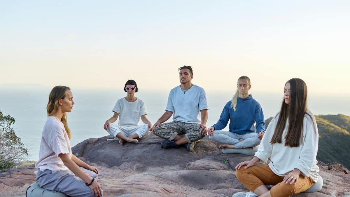 Five adults meditating outdoors on a mountain top during sunrise, promoting relaxation and mindfulness.