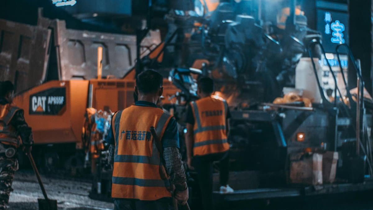 Workers in safety vests operate heavy machinery at a night construction site.