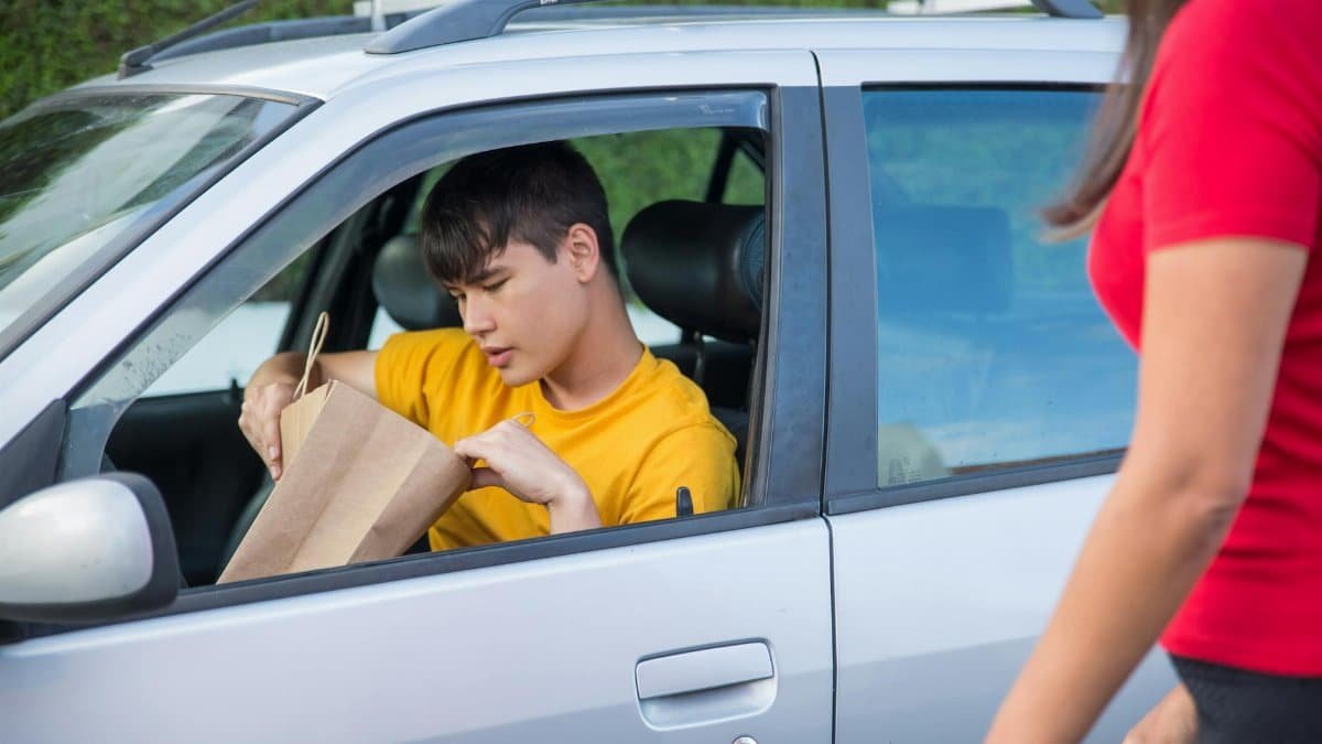 A driver in a yellow shirt delivers a paper bag order to a customer beside a car.