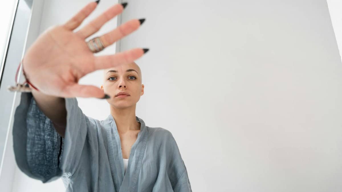 Bald woman in a robe making a stop gesture indoors, symbolizing resistance or caution.