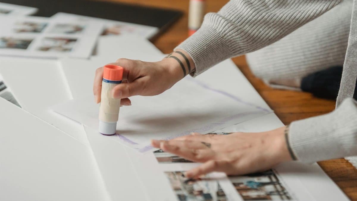Crop anonymous person in warm gray wear applying glue stick on papers and arranging photo album on floor