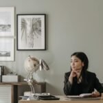 Asian woman in black blazer sitting at a desk in stylish home office, deep in thought.