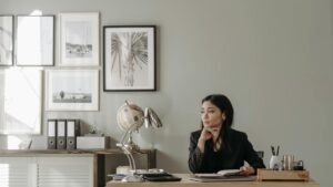 Asian woman in black blazer sitting at a desk in stylish home office, deep in thought.