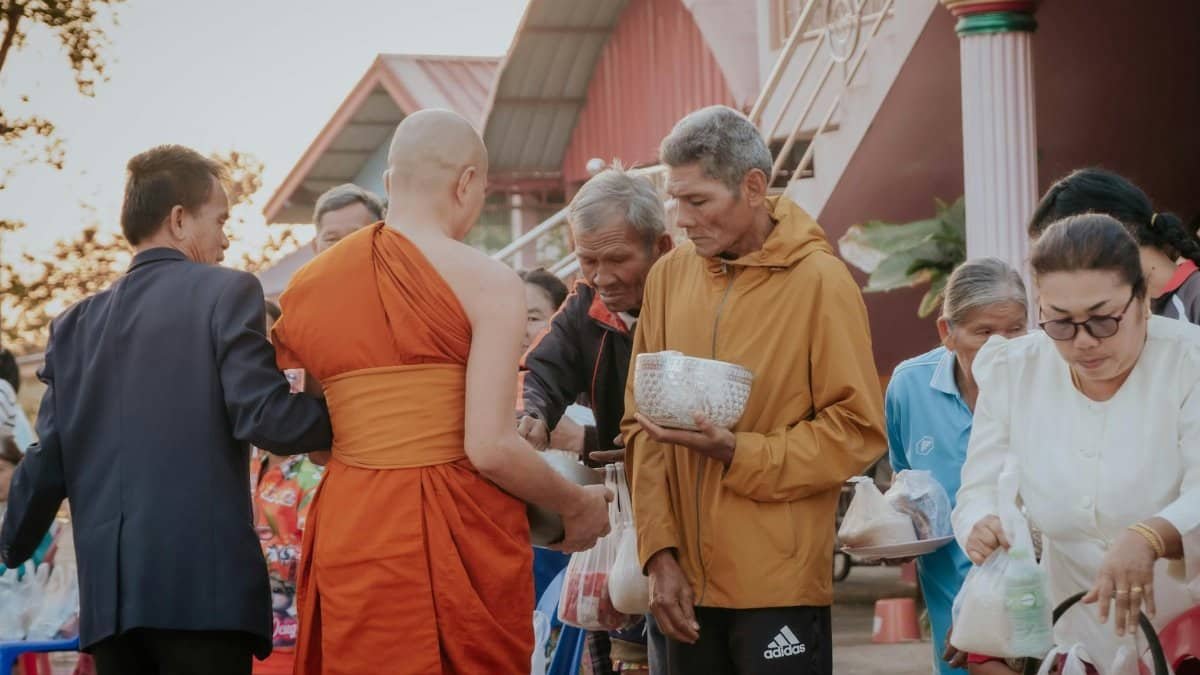 A group of people giving alms to Buddhist monks during a religious ceremony outdoors, in traditional setting.