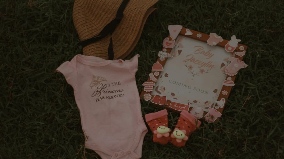 Flat lay of baby arrival announcement with pink onesie and accessories on grass.