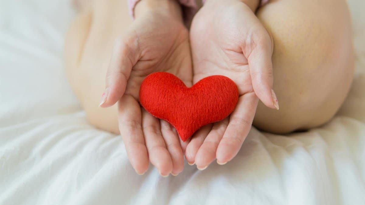 Unrecognizable female sitting with bare legs on white sheet with small red heart in hands in light room in daytime