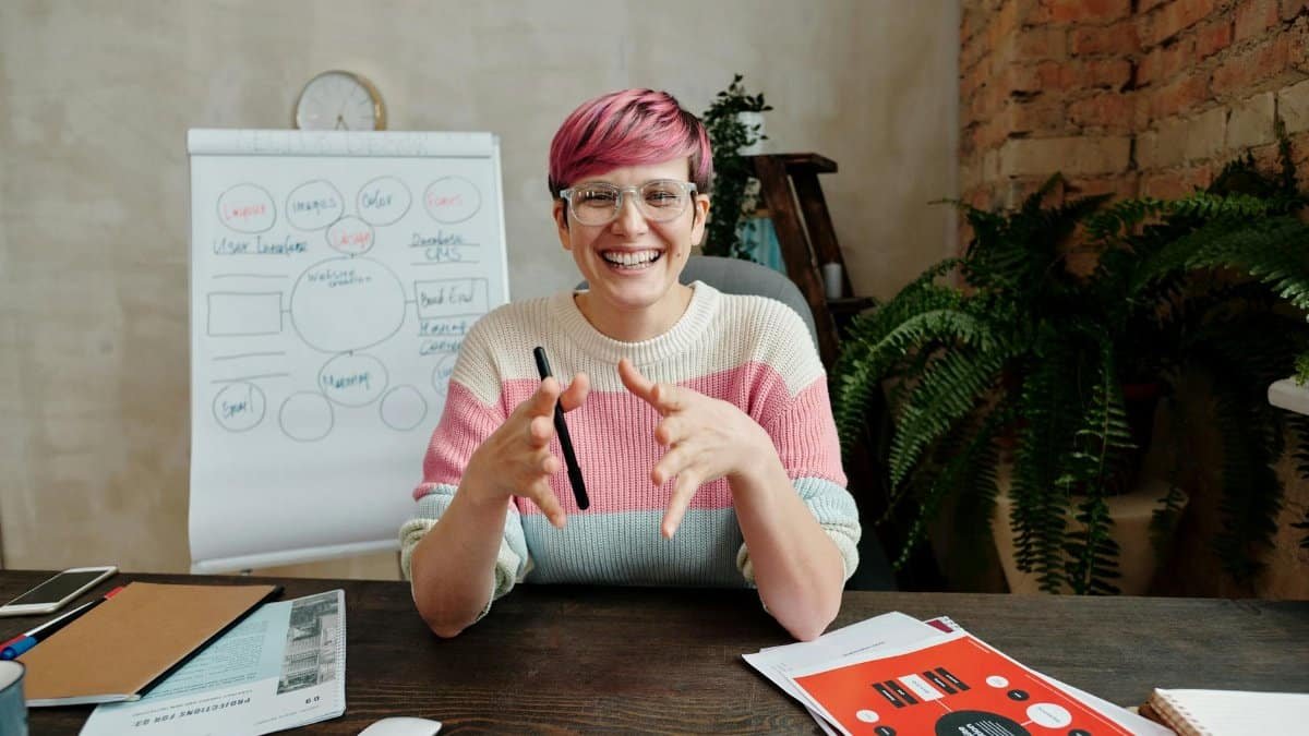 Smiling woman with pink hair in casual sweater leading an office meeting with visual aids.