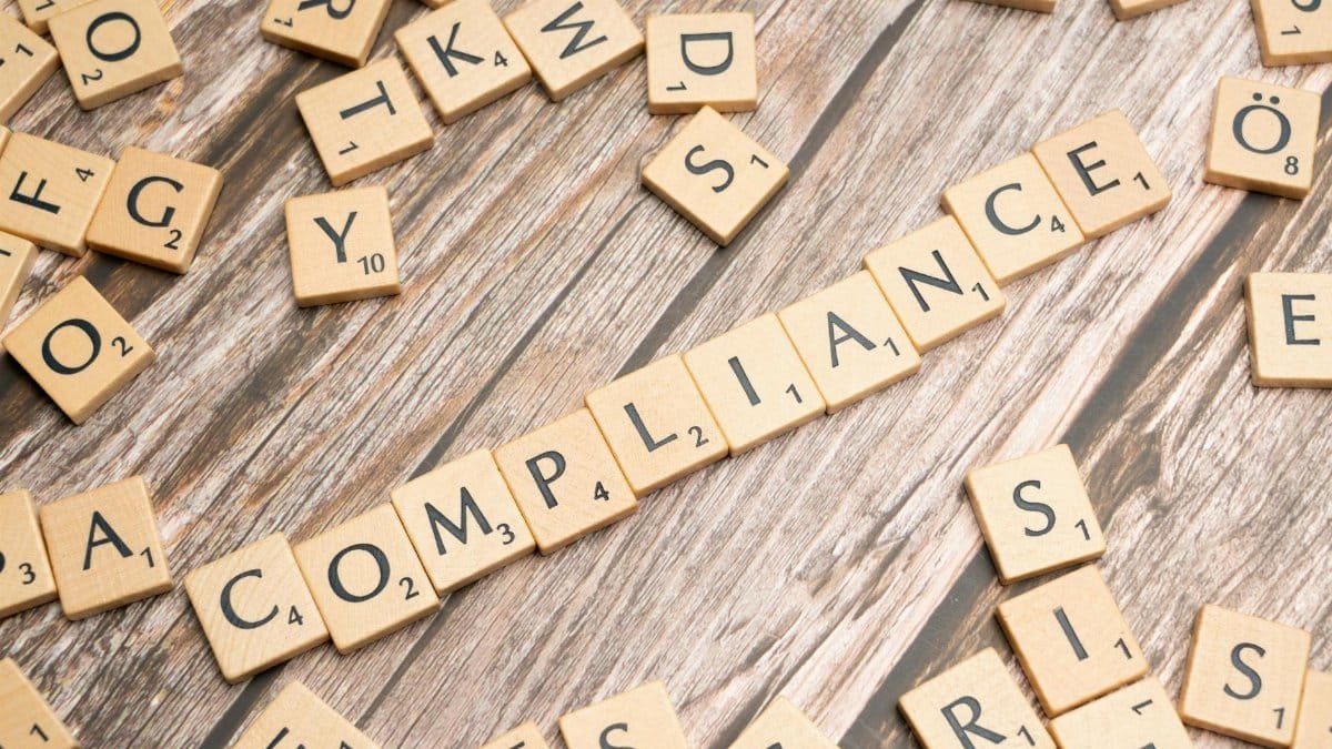 Wooden letter tiles forming the word 'COMPLIANCE' on a rustic wooden background.
