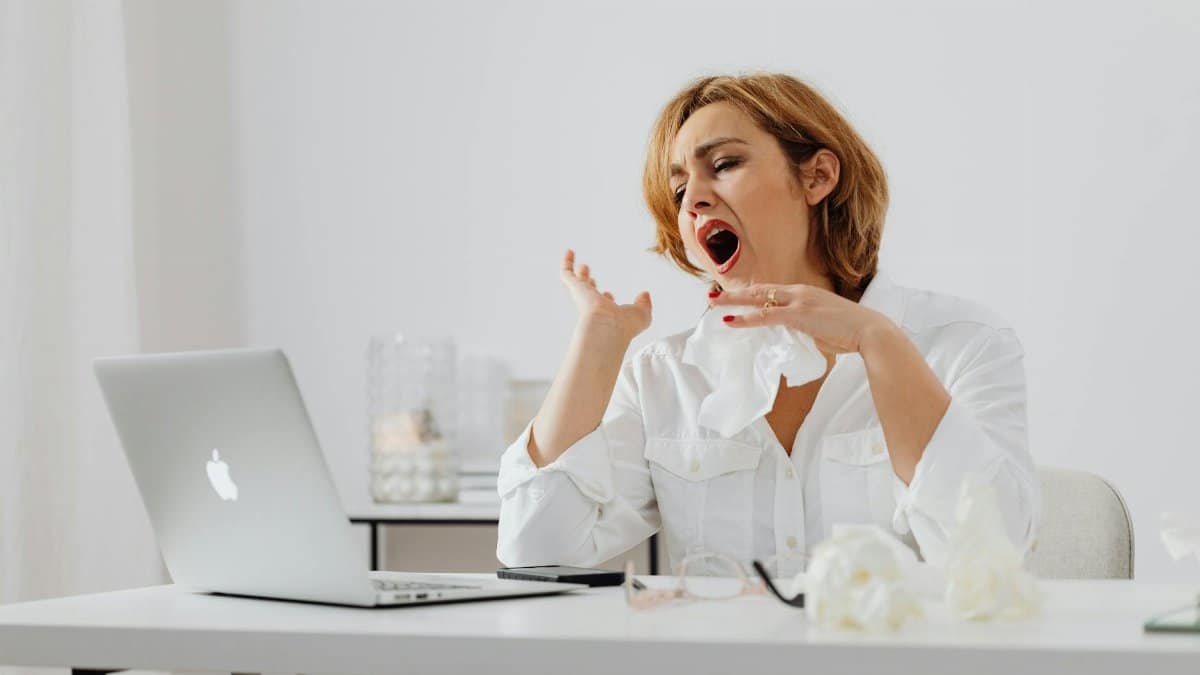 A woman in white yawning while working at a desk, indicating fatigue.