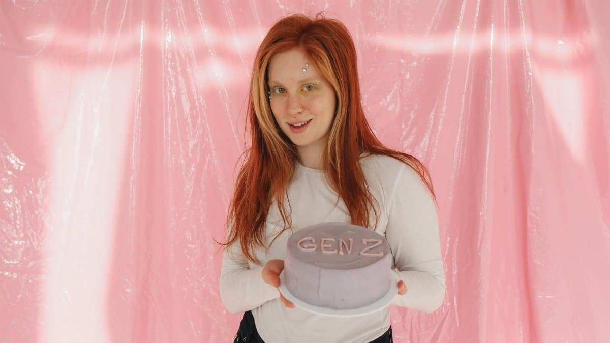 Smiling redhead woman in a white top holding a 'Gen Z' cake against a pink background.