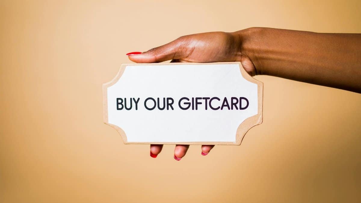 Close-up of a hand holding a 'Buy Our Giftcard' sign on a brown backdrop.