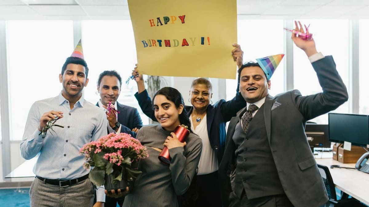 Happy colleagues celebrating a birthday in an office with hats and balloons.