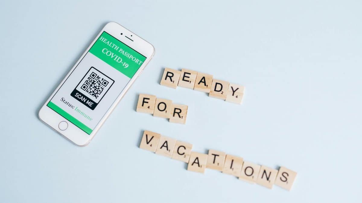 Smartphone displaying COVID-19 health passport next to Scrabble letters spelling 'Ready for Vacations' on a pastel background.