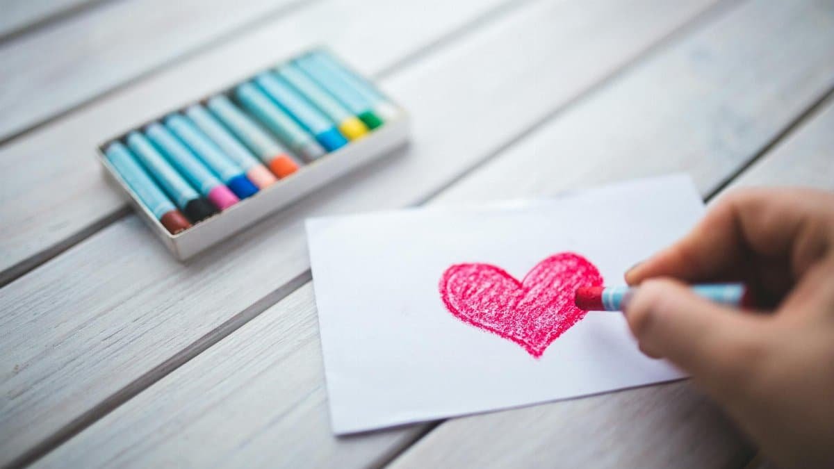 A close-up of a hand using pastel crayons to draw a red heart on paper.
