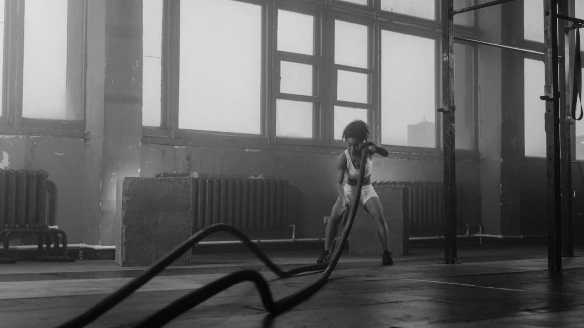 Black woman in gym performing an intense battle ropes workout, highlighting strength and fitness.